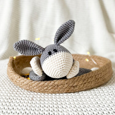 Grey and cream crocheted baby snuggle donkey with long ears in a woven tray on a cream knit blanket