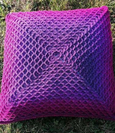Quadratischer gehäkelter Kissenbezug mit pink‑violettem Farbverlauf und offenem Netzmuster auf Gras
