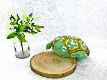 Crocheted green amigurumi turtle with African flower granny motif shell on a wood slice next to a glass vase of small white flowers