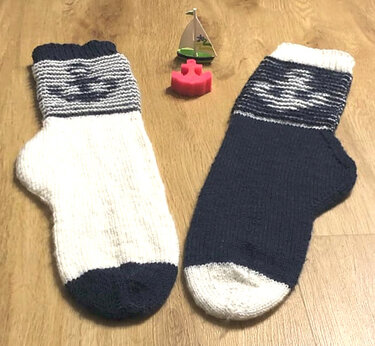 Zwei handgestrickte Socken in Blau und Weiß mit Ankermotiv am Schaft auf Holzboden, dazwischen ein kleines Spielzeugboot.