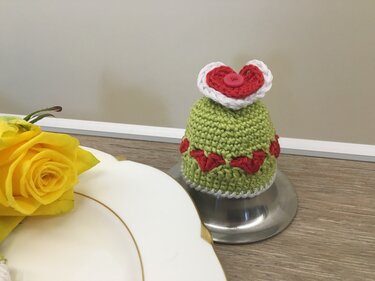 Hellgrüner gehäkelter Eierwärmer mit roten Herzapplikationen und Knopf auf Metallständer neben gelber Rose und Teller