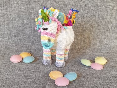 Gehäkeltes Einhorn-Amigurumi in Pastellfarben mit offenem Fach und Süßigkeiten auf grauem Hintergrund