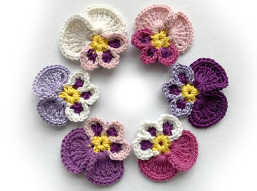 Sechs gehäkelte Stiefmütterchen in Weiß, Rosa und Lila mit gelber Mitte, kreisförmig auf weißem Hintergrund angeordnet.