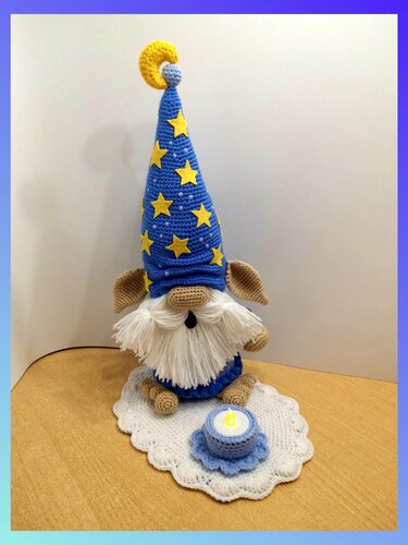Gehäkelter Amigurumi-Wichtel mit blauem Sternenhut, weißem Bart und Kerze auf weißem Deckchen