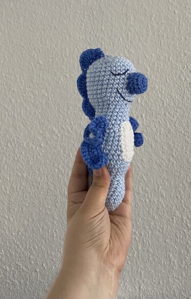 Hand hält hellblaues gehäkeltes Seepferdchen-Amigurumi mit dunkelblauen Flossen und weißem Bauch