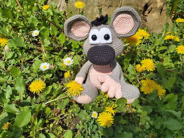 Gehäkelte Amigurumi-Maus in Grau mit rosa Ohren und dunkler Schnauze, sitzend zwischen Löwenzahn und Gänseblümchen