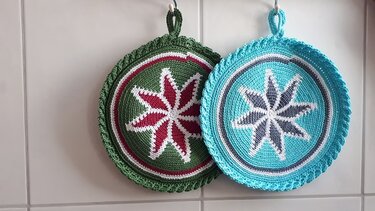 Zwei runde Topflappen, grün mit rotem Stern und türkis mit grauem Stern, aufgehängt an einer hellen Fliesenwand.