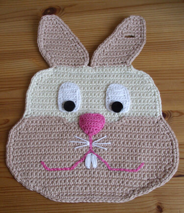 Gehäkelter Topflappen in Hasenform mit rosa Nase auf Holzuntergrund