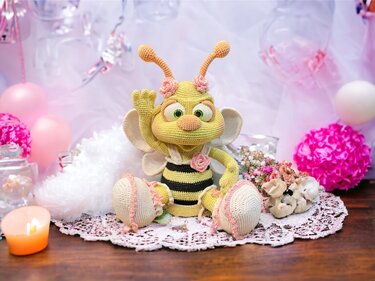 Gehäkelte gelb-schwarze Amigurumi-Biene mit Flügeln und Rosen, sitzend auf einem Spitzen-Deckchen, umgeben von pastellfarbener Dekoration