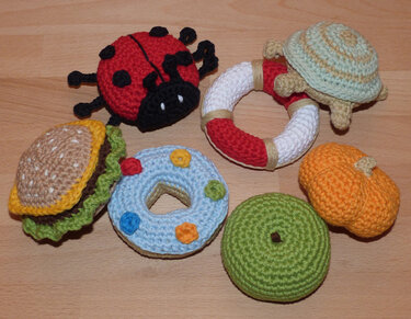 Mehrere gehäkelte Babyspielzeuge (Marienkäfer, Rasselring, Donut, Hamburger, Kürbis) auf hellem Holzuntergrund