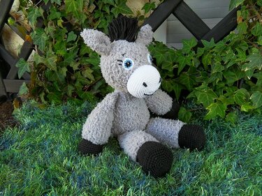 grauer gestrickter Amigurumi-Esel sitzend mit schwarzen Hufen