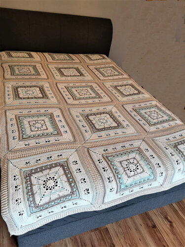 Gehäkelte Decke aus quadratischen Motiven in Creme, Beige, Grau und zarten Pastelltönen auf einem Bett