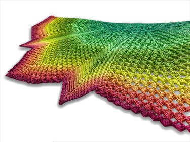 Close-up of a triangular crochet shawl in rainbow gradient yarn showing open lace mesh and pointed scalloped edges