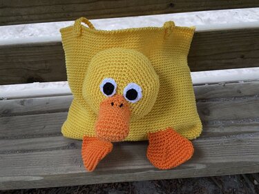 Gelbe Häkel-Tasche mit aufgesetztem Kopf, angenähtem orangefarbenem Schnabel und Fußapplikationen sowie zwei Schlaufen
