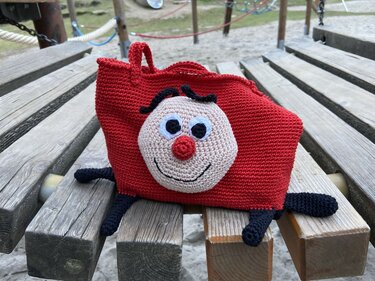 Rote gehäkelte Tasche mit aufgesetztem Gesicht, großen Augen, roter Nase und schwarzen Beinen auf einer Holzbank