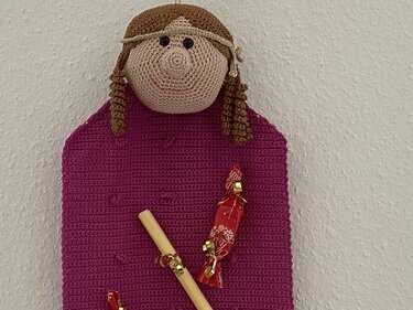 Gehäkelte Puppe mit braunen Zöpfen und magentafarbenem Körper an weißer Wand, zwei eingesteckte Bonbons