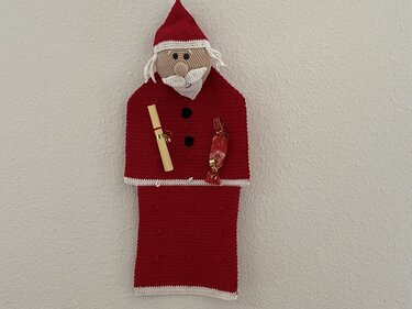Gehäkelter Weihnachtsmann-Wandbeutel in Rot mit Mütze und zwei Taschen, an einer weißen Wand hängend