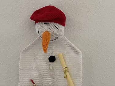Gehäkelter weißer Schneemann-Wandbehang mit roter Mütze und orangefarbener Nase.