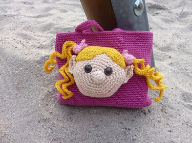 Pinkfarbene handgehäkelte Tasche mit appliziertem Mädchenkopf, gelben Locken und rosa Schleifen auf sandigem Untergrund
