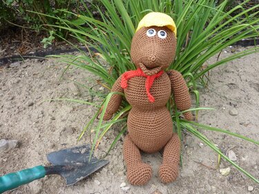 Braunes Maulwurf-Amigurumi mit gelbem Helm und rotem Halstuch im Sand, links eine Gartenschaufel