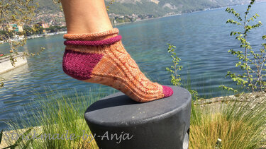 Handgestrickte kurze Socke in Orange und Magenta mit Zopfmuster auf einem Fuß vor einem See und Bergen.