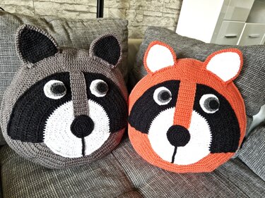 Zwei runde gehäkelte Waschbär-Kissen, grau und orange, auf einem Sofa