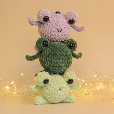 Drei gestapelte gehäkelte Frosch-Amigurumi in Pastell- und Grüntönen vor neutralem Hintergrund mit Lichterkette