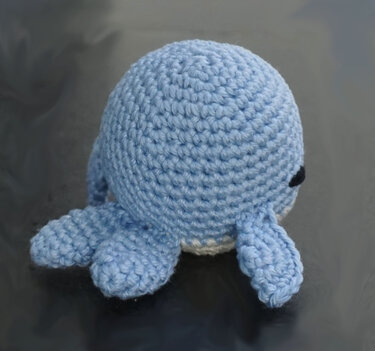 Hand-crocheted light blue amigurumi octopus with round body and short tentacles on a dark background