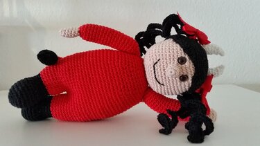 gehäkelte Amigurumi-Puppe liegend, roter Overall, schwarze Locken, rote Schleifen, kleine weiße Hörner