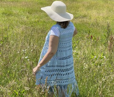 Rear view of a sleeveless long crochet vest with alternating openwork panels and fringe worn over a white t-shirt in a grassy field