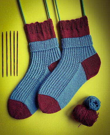 Paar gestrickter Damensocken in Blau und Burgunder mit Garnknäueln und Rundstricknadeln vor gelbem Hintergrund
