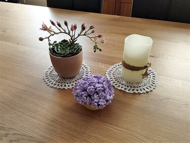 Sukkulente, Kerze und lila Häkelblüte auf weißen Spitzenuntersetzern auf Holztisch.