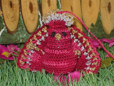 Gehäkelter Amigurumi-Vogel in Magenta mit goldenen und silbernen Garnakzenten auf grünem Gras