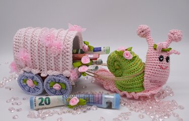 Gehäkelte rosa Schnecke zieht einen blütenverzierten Wagen mit gerollten Geldscheinen.