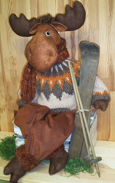 Stoffelch mit braunem Geweih, gemustertem Strickpullover, Schal und braunen Hosen, sitzend neben rustikalen Holzskiern.