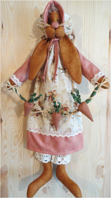 Genähter Stoffhase mit rosa Kapuze, geblümtem Kleid und karierten Stoffkarotten an Holzwand