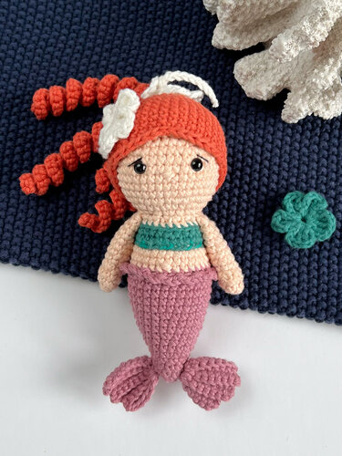 Amigurumi‑Meerjungfrau mit rötlich‑orangen Locken, grünem Oberteil und mauvefarbener Flosse auf dunkelblauem Häkelgrund