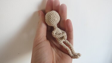 Hand holding a small beige crocheted amigurumi doll body with round head and long legs