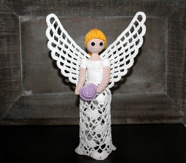 Gehäkelter Amigurumi-Engel in Weiß mit gelben Haaren, großen, durchbrochenen Flügeln und einem lila Herz in den Händen vor dunklem Holzrahmen.