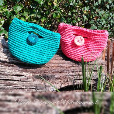 Zwei handgehäkelte Taschen in Türkis und Pink mit runden Verzierungen auf einem Holzstamm vor grünen Blättern.