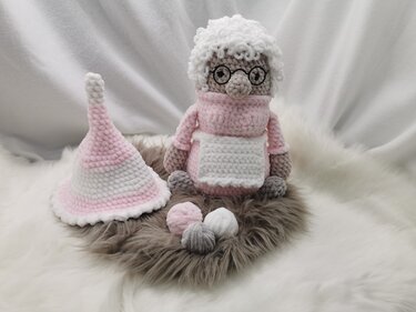Gehäkelte Oma-Puppe mit rosa Kleidung, Brille, weißem lockigem Haar und weißer Schürze, sitzend neben einer rosa-weißen gestreiften Wichtelmütze und drei kleinen gehäkelten Kugeln auf flauschigem Untergrund.