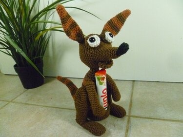 Braunes gehäkeltes Amigurumi-Tier mit gestreiften Ohren und großen Augen, sitzt auf Fliesen neben einer grünen Pflanze.