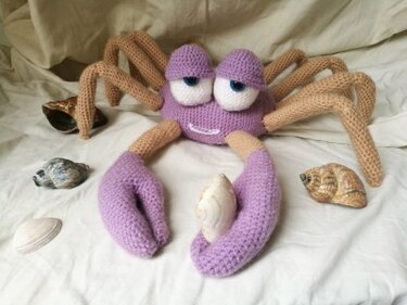 Gehäkelte lila-beige Krabbe mit großen Augen und Muscheln auf hellem Stoff