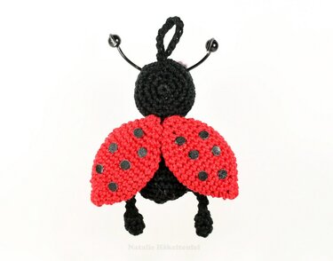 Nahaufnahme eines gehäkelten Marienkäfers in Rot und Schwarz mit Fühlern und Aufhängeschlaufe vor weißem Hintergrund.