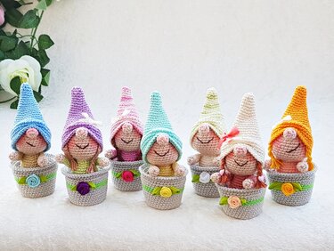 Sieben gehäkelte Amigurumi-Wichtel mit Pastellmützen in grauen Töpfen, dekoriert mit Bändern und Rosen.