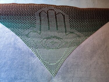 Gehäkeltes Dreieckstuch im Filetmuster mit zentralem Handmotiv und Farbverlauf von Braun zu Grün, ausgebreitet auf hellem Untergrund