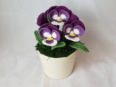 Drei gehäkelte Stiefmütterchen in Lila, Weiß und Gelb in einem kleinen beigen Blumentopf mit grünem Moos