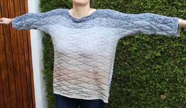 Weit geschnittener gestrickter Damenpullover mit Wellenmuster und Farbverlauf von blau-grau zu beige, Arme seitlich ausgestreckt