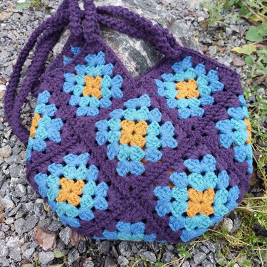 Boho-Umhängetasche aus gehäkelten Granny-Squares in Violett, Hellblau und Senfgelb, auf Kies liegend
