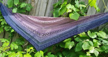 Gehäkeltes Dreieckstuch mit Farbverlauf von Blau zu Rosa und durchbrochenem Muster vor einem Holzzaun mit Blättern
